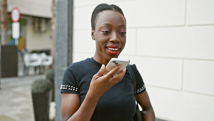 Smiling african american woman joyously sending a voice message over her smartphone, standing on a...