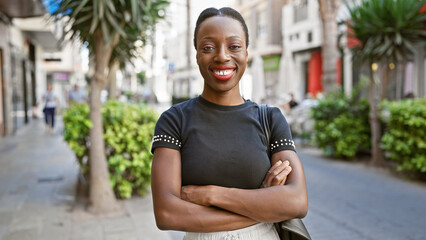 Joyful african american woman standing with arms crossed on a city street, displaying a beautiful smiling expression of happiness - obrazy, fototapety, plakaty