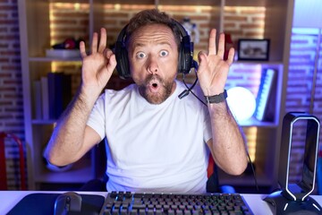 Middle age man with beard playing video games wearing headphones looking surprised and shocked...