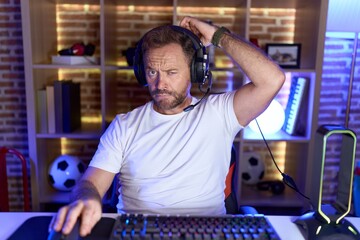 Middle age man with beard playing video games wearing headphones confuse and wondering about question. uncertain with doubt, thinking with hand on head. pensive concept.