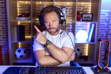 Middle age man with beard playing video games wearing headphones pointing with hand finger to the side showing advertisement, serious and calm face