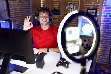 Young hispanic man playing video games recording with smartphone doing ok sign with fingers,...
