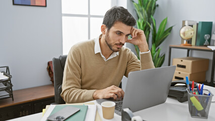 Focused hispanic man with beard working in a modern office setting, reflecting concentration and...