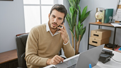 Hispanic man with beard wearing sweater talking on phone while working on laptop in modern office.