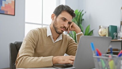 Handsome hispanic man with beard in office using laptop, portrayed in thoughtfully casual attire.