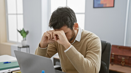A stressed young hispanic man with a beard is showing frustration or headache in a modern office setting.