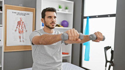 Handsome young hispanic man exercising with dumbbells in a rehabilitation clinic room.