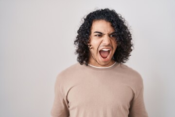 Fototapeta na wymiar Hispanic man with curly hair standing over white background angry and mad screaming frustrated and furious, shouting with anger. rage and aggressive concept.