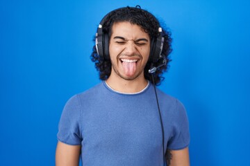 Hispanic man with curly hair listening to music using headphones sticking tongue out happy with funny expression. emotion concept.