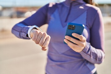Young beautiful hispanic woman wearing sportswear using smartphone looking stopwatch at street