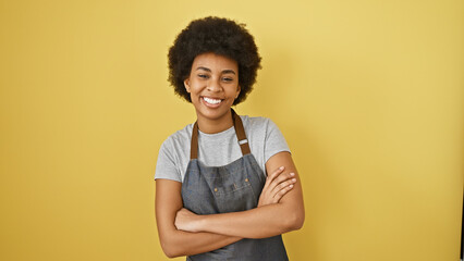 Obraz na płótnie Canvas Portrait of a smiling woman with curly hair and crossed arms wearing an apron against a yellow background.