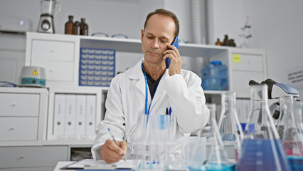 Serious middle-aged man, a scientist absorbed in writing his research report, while talking on a smartphone in the lab.