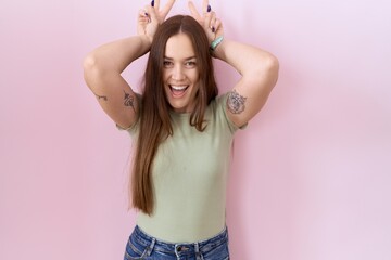 Beautiful brunette woman standing over pink background posing funny and crazy with fingers on head...