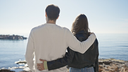 A couple embraces while gazing at the ocean from a sunny seaside, symbolizing love and togetherness in nature.