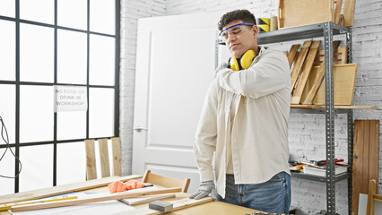 A young man with a beard experiences shoulder pain in a well-organized carpentry workshop indoor.