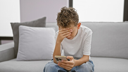 Adorable blond boy, stressed and upset, sitting on sofa at home using smartphone. worried child typing a message, showing serious expression amidst technology stress.