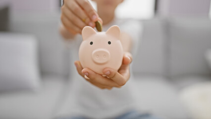 Adorable blond boy comfortably inserting savings into his piggy bank while relaxing on the sofa at home, conveying the lessons of finance and investment in a child's lifestyle.