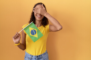 Young hispanic woman holding brazil flag smiling and laughing with hand on face covering eyes for surprise. blind concept.