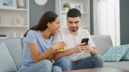 Obraz premium Beautiful couple together in love, confidently smiling, texting on smartphone over coffee on living room sofa at home