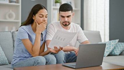 Stressed beautiful couple at home, reading upsetting document on laptop, worrying together in their living room