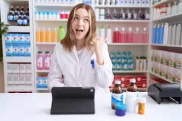 Young caucasian woman working at pharmacy drugstore using tablet smiling with happy face looking and pointing to the side with thumb up.