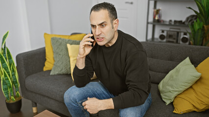 A hispanic man looking concerned while talking on a smartphone in a modern living room setting