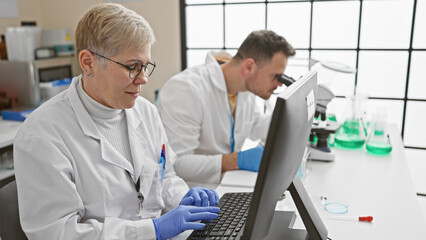 A woman and a man in lab coats work diligently in a bright laboratory, focusing on computer...