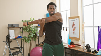 A young african american man stretching his arm in a rehabilitation clinic's therapy room, indicating exercise and healthcare.