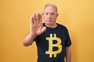 Senior man with grey hair wearing bitcoin t shirt doing stop sing with palm of the hand. warning...