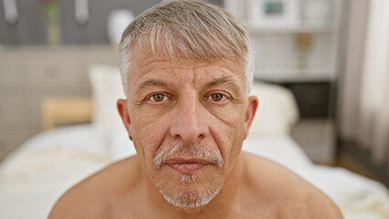 A shirtless, mature man with gray hair sits in a bedroom, giving a detailed, close-up view of...