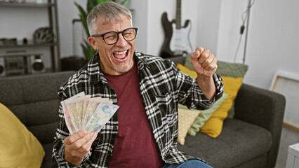 Excited middle-aged man holding british pounds celebrates indoors with a guitar in the background.