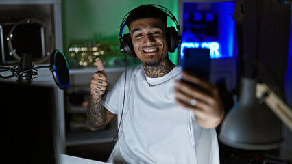 Smiling, tattooed young latin man, a music studio maestro, snapping a vibrant selfie with his smartphone amidst the recording session, donning his headphones. a solo night cap in his artistic haven. - obrazy, fototapety, plakaty