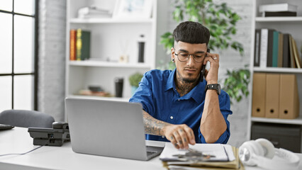 Handsome young latin man, a serious business worker engrossed in online work on his laptop, deeply focused in a heated phone conversation on his smartphone at the office.