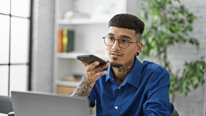 Focused young latin man, tattooed business worker, captured in successful office portray, sending voice message via smartphone at his indoor workplace