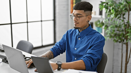 Concentrated young latin man at work, elegantly dressed, business professional operating a laptop and touchpad in his office environment. a serious, focused worker managing success, indoors.