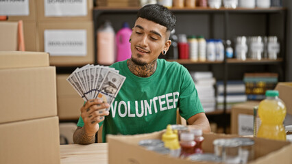 Handsome, tattooed young latin man, smiling confidently, volunteering at the charity center, holding dollars for donations in his hands
