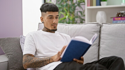 Relaxing at home, a young tattooed latin man comfortably absorbs knowledge reading literature from...