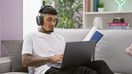 Young latin man using laptop and headphones reading book at home