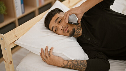 Handsome, tattooed young latin man in pyjamas relaxing in comfortable bed, having a serious talk on...