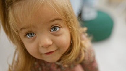 Close-up of a curious little girl with blonde hair and blue eyes indoors, evoking a sense of...