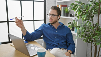 Young hispanic man business worker using laptop and earphones holding pen at the office