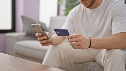 Young hispanic man shopping with smartphone and credit card sitting on sofa at home