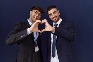 Two hispanic business men wearing business clothes smiling in love doing heart symbol shape with hands. romantic concept.