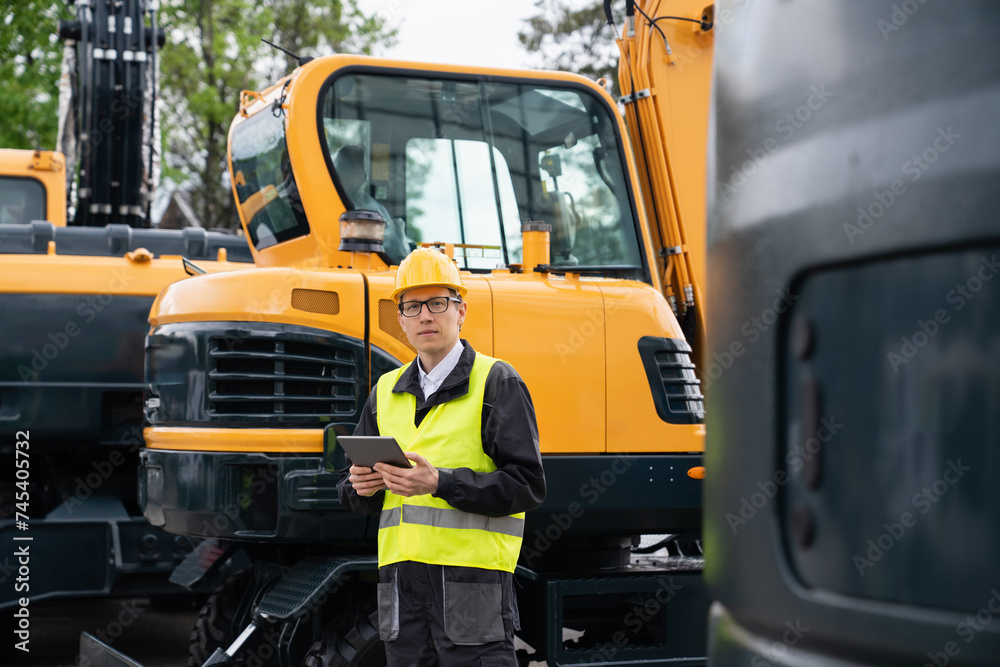 Poster Engineer in a helmet with a digital tablet stands next to construction excavators..