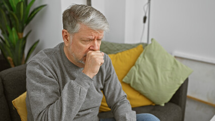 A thoughtful mature man sits indoors, pondering deeply on a couch in a cozy living room.
