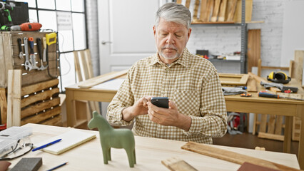 Mature man using smartphone in a woodworking workshop surrounded by tools and workbench.
