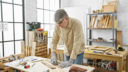 Grey-haired man measuring wood in a bright carpentry workshop.