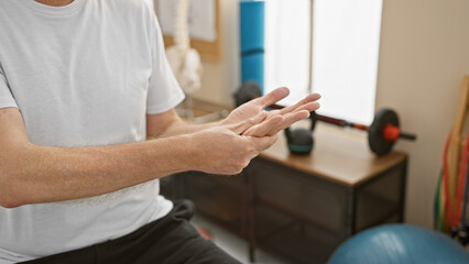 A middle-aged man examines his wrist in a rehabilitation clinic's setup with exercise equipment.