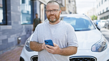 Handsome middle-aged caucasian man, with a confident beard, enjoy standing by his car on a sunlit city street. he's casually texting on his smartphone displaying a happy smile.