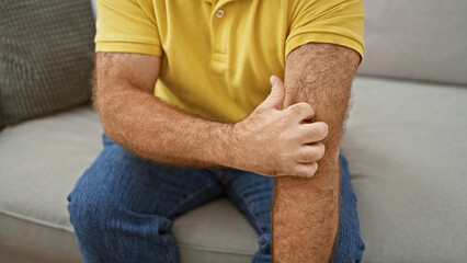 Handsome middle-aged caucasian man sitting on sofa at home, scratching irritated skin on arm. itchy dermatitis rash causing discomfort. health care needed for allergic reaction. - obrazy, fototapety, plakaty